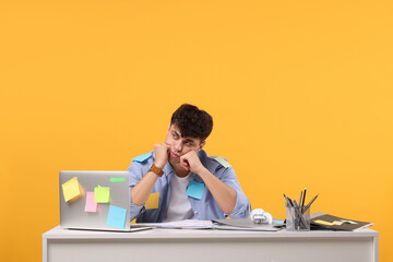 Tired young man working at white table on orange background. Deadline concept