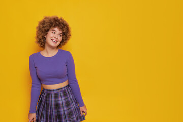 Curly girl in violet colour longsleeve top stands on yellow background and smiles, holds hands down, happy moments concept, copy space