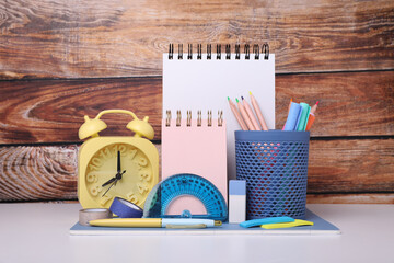 Different school stationery and alarm clock on white table near wooden wall. Back to school - obrazy, fototapety, plakaty