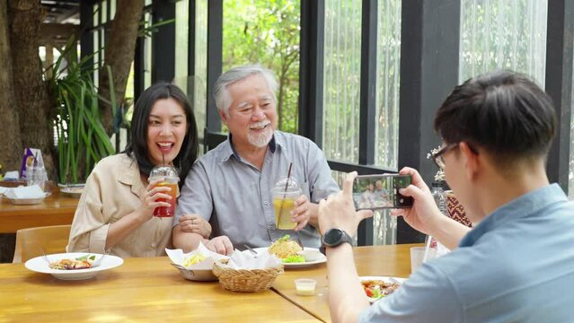 Asian man using mobile phone taking picture his wife and elderly father during having lunch eating food together at restaurant on summer vacation. Family relationship old people health care concept.