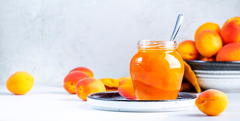 Apricot jam or confiture in glass jar with spoon, fresh fruit on gray kitchen table background