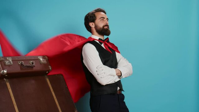 Hotel worker with red mantle to help guests with trolley bags, pretending to be fantasy cartoon superhero character on camera. Bellhop acting like comic superhuman with cape against background.