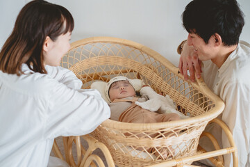 春の日中、ゆりかごで眠る女の子の赤ん坊を囲んで微笑む日本人の夫婦 - obrazy, fototapety, plakaty