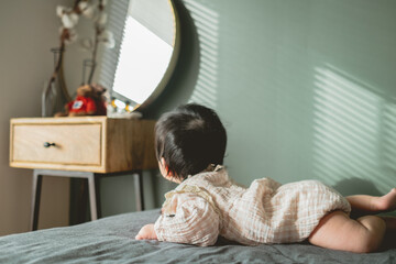 秋の昼間、ベッドの上でうつ伏せでドレッサーの方を見つめるアジア人のロンパースを着た赤ん坊の後ろ姿 - obrazy, fototapety, plakaty