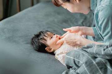 春の昼間、ベッドで横たわる日本人の赤ちゃんとほっぺに触れてあやす母親