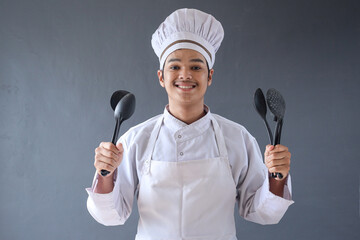 Young Asian male chef in white apron holding utensils isolated on grey background