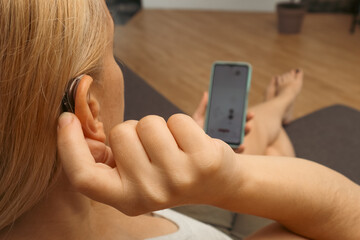 Hearing impaired woman working with smartphone. Close up of blonde woman wearing hearing aid on ear and pressing button while using smartphone at home.