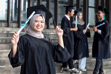 A young Asian muslim girl graduate in hijab holding diploma standing against the background of...