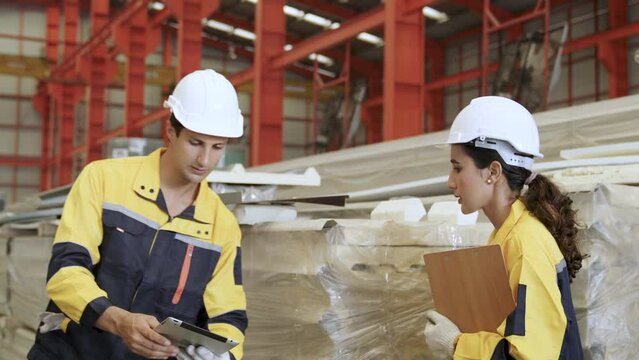 In a factory, male and female engineers wearing industrial helmets stand holding laptops and documents. Review the materials produced to show teamwork and production.