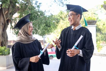 Two Asian graduate students chatting and laughing while holding certificate on graduation day