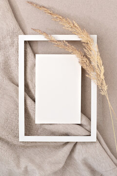 Blank paper card mock up in empty picture frame border, dried meadow grass on neutral beige fabric background. Aesthetic wedding invitation, congratulation postcard design, beauty brand template