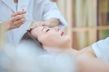 아시아 한국의 젊은 한국 여성 모델이 스파 또는 피부관리전문점 또는 피부과 병원에서 침대에 누워서 앰플 스포이드를 들고 있는 전문피부 관리사에게 피부 관리를 받고 있다.