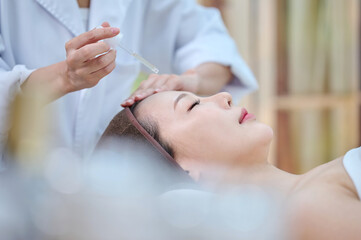 Obraz na płótnie Canvas 아시아 한국의 젊은 한국 여성 모델이 스파 또는 피부관리전문점 또는 피부과 병원에서 침대에 누워서 앰플 스포이드를 들고 있는 전문피부 관리사에게 피부 관리를 받고 있다.