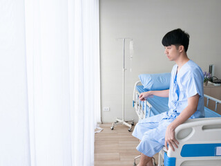 Asian patient man sitting with absent minded when he know about disease diagnosis in hospital bed near curtain window in the private room while wearing inpatient uniform
