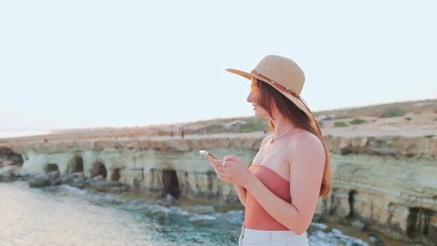 Side-view portrait of happy cute young woman taking picture of beautiful sunset and sending messages texting online using smartphone smiling. Young attractive girl wearing hat enjoying vacation.
