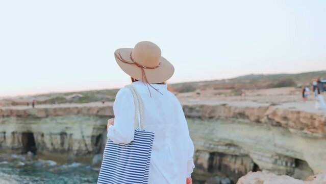 Back view portrait of joyful young Caucasian woman standing on the cliff by the seaside wearing hat enjoying beautiful views of ocean. Vacation of attractive girl relaxing looking at sea having fun.