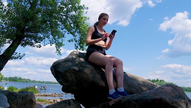 Positive charming athletic fit woman sitting on rock by riverbank , networking and online messaging using smartphone while relaxing in summer nature after intense sports training.