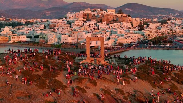Aerial view of the famous Portara monument and Naxos town with tourists enjoying the sight during soft summer sunset light, Cyclades, Greece