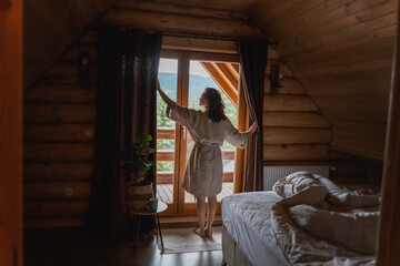 Fototapeta na wymiar Young caucasian woman opening curtains in a wooden chalet cabin in the mountains.