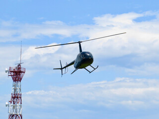 Black helicopter in flight. A small helicopter flies past a cell tower against a blue sky and white clouds.