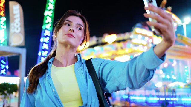 Beautiful charming young woman taking pictures of herself using smartphone standing at amusement park at nighttime. Attractive adult Caucasian cute girl taking selfies. Camera concept.
