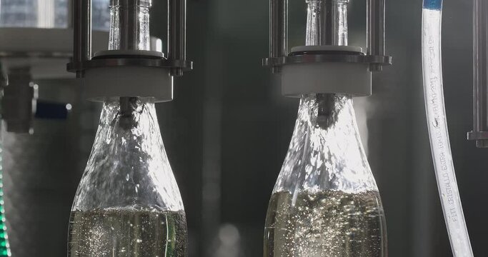 Close-up shot of filling white wine by the machine into bottles made of transparent glass. White wine bottling production line. Slow motion
