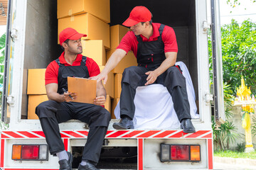 Multiethnic employees in uniform work professional teamwork check property lists sit in delivery cargo truck full of cardboard box background, moving house workers wait to move customer's furniture