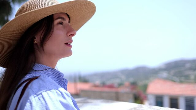 Side view of beautiful Caucasian woman in Europe for summer vacation standing on balcony, enjoying mountain views. Attractive inspired happy tourist girl with hat looking at sights.