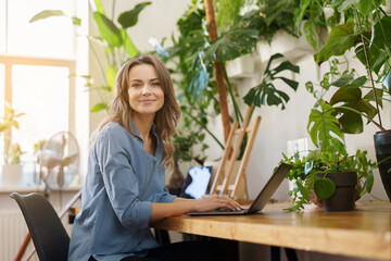 A charming smiling freelance woman in a blue shirt working on her laptop in a plant-filled room