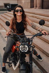 Fototapeta na wymiar Stylish brunette in jeans and a tank top, with sunglasses and a tattoo on her arm, poses on her retro motorcycle on an old cobblestone street in Europe