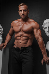 Fototapeta na wymiar An attractive, muscular man in black jeans poses alongside ancient Greek sculpture on a black background, creating a visually striking contrast between the modern and the classical