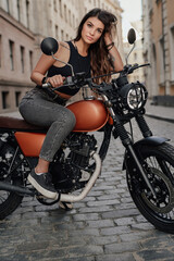 Fototapeta na wymiar In the old-world charm of a paved alleyway in Europe, a beautiful young woman with dark hair stands proudly next to her vintage motorcycle
