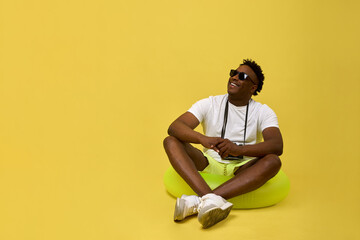 Fototapeta na wymiar Handsome African young man in white T-shirt crouched on a lifebuoy, smiling, looking up. On a yellow background, contrasting African guy in sunglasses rests sitting in an inflatable swimming circle