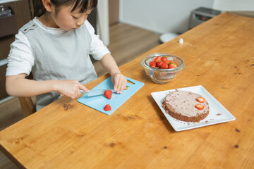 春の日中に屋内のテーブルでケーキに乗せる苺を切る日本人の女子 - obrazy, fototapety, plakaty