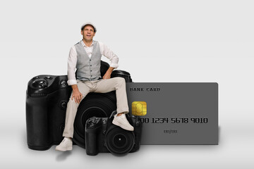 Young successful man sitting on modern camera near credit card isolated on gray background, Driven and prosperous man confidently positioned on modern camera, embodying success and financial prowess.