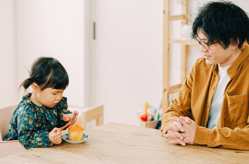 Fototapeta premium 冬の昼間、座ってフォークでカップケーキを食べる日本人の子どもと見つめる父