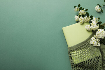 Book in string bag and flowers on green background, space for text