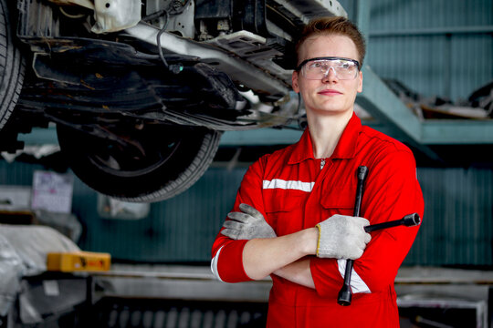 Portrait of smiling mechanic in red uniform standing arms crossed near lifted vehicle, auto mechanic check wheel at garage, technician check, maintenance customer car automobile at repair service shop