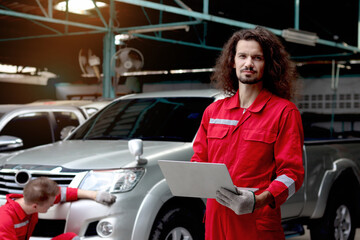 Mechanic in red uniform holding laptop computer during coworker repairing paint car after accident, auto mechanic technician team doing maintenance customer automobile vehicle at garage service shop.