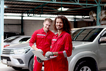Two handsome mechanic men in red uniform holding laptop computer during discussion together at vehicle garage, coworker technician team repairing maintenance customer car automobile at service shop.
