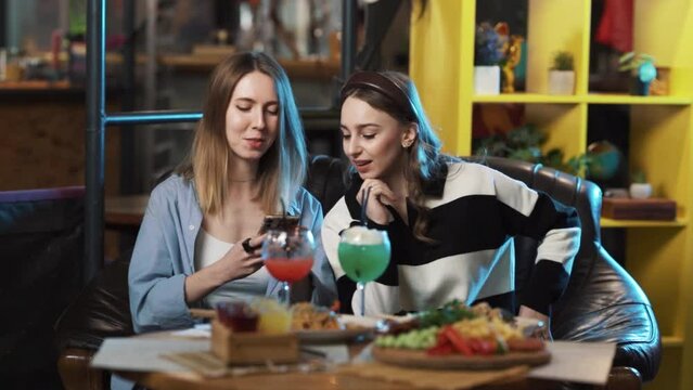 The girls are sitting at a restaurant, in high spirits, talking about something, laughing. One of the girls holds a phone in her hands and shows something to the other.