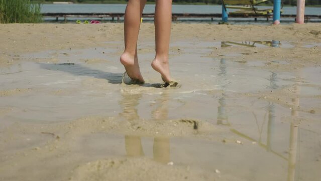 Kid plays on the beach near lake. Happy little girl in a swimsuit jumping and playing in sand in puddle. Close up of legs