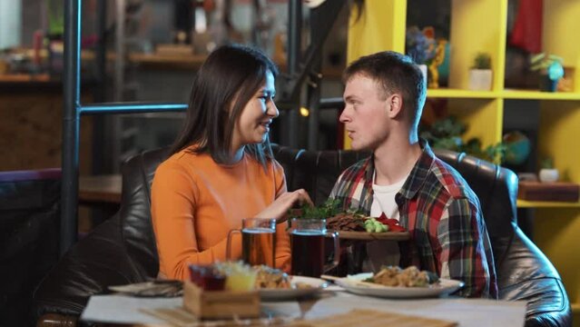 The couple is sitting in a restaurant, they have a romantic atmosphere, and they have ordered some dishes and drinks. The girl is feeding the guy with her hands.