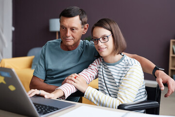 Portrait of smiling teenage girl with disability using laptop computer at home with father helping, copy space