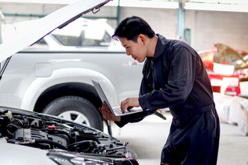 Asian mechanic man in uniform holding laptop computer during working with engine vehicle, auto mechanic technician inspecting maintenance customer car automobile at garage car repair service workshop.