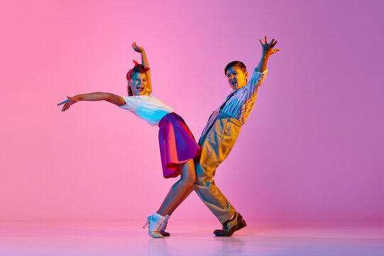 Talented, artisitc children, boy and girl in retro clothes dancing lindy hop against pink studio background in neon light. Concept of childhood, hobby, active lifestyle, performance, art, fashion