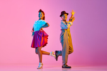 Emotional, beautiful children, boy and girl in retro costumes dancing lindy hop against pink studio background in neon light. Concept of childhood, hobby, active lifestyle, performance, art, fashion