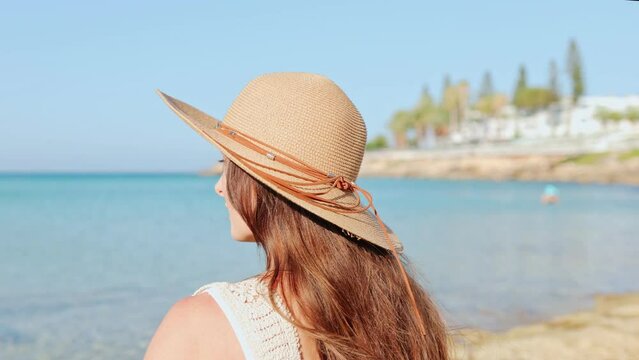 Back view portrait of beautiful young Caucasian woman enjoying summer vacation sitting at beach sunbathing. Attractive cute girl wearing hat spending time by the sea. Relaxation concept.