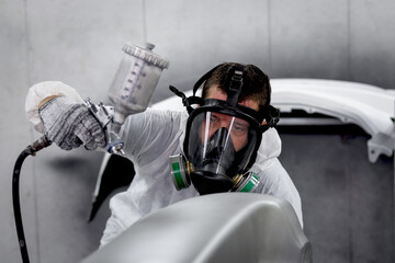 Mechanic man in white costume cloth wearing chemical protective mask while working with painting tool, auto mechanic painting a car, fixing repairing vehicle at garage automobile repair service shop.