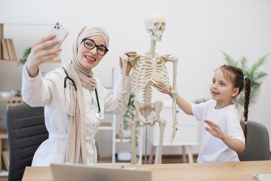 Physician in hijab taking self-picture with kid in office
